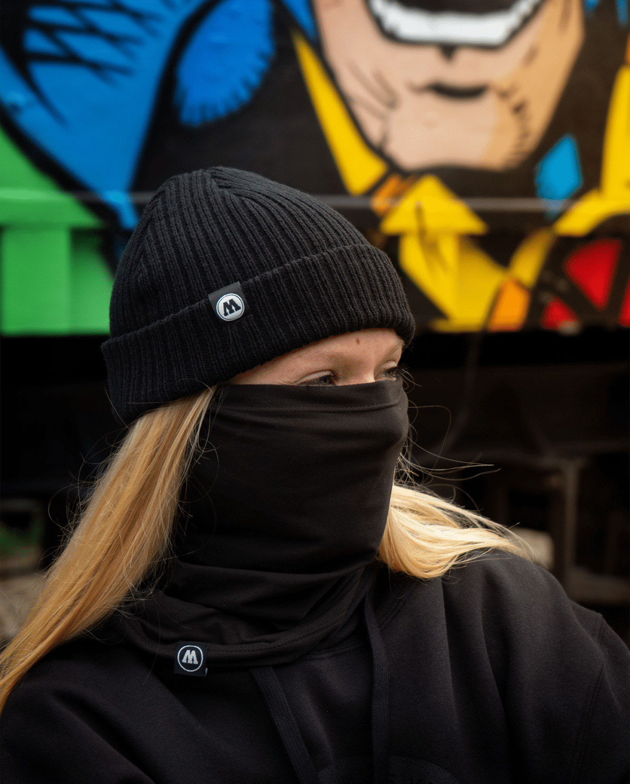 Person wearing a black Molotow beanie and neck warmer in front of a graffiti wall